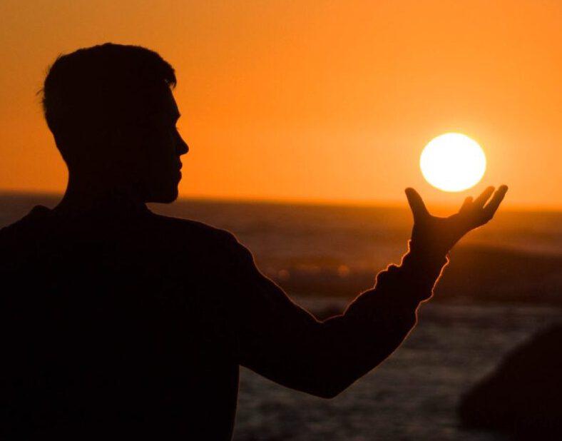 Mann sitzt vor Sonnenuntergang am Meer und hält die Sonne