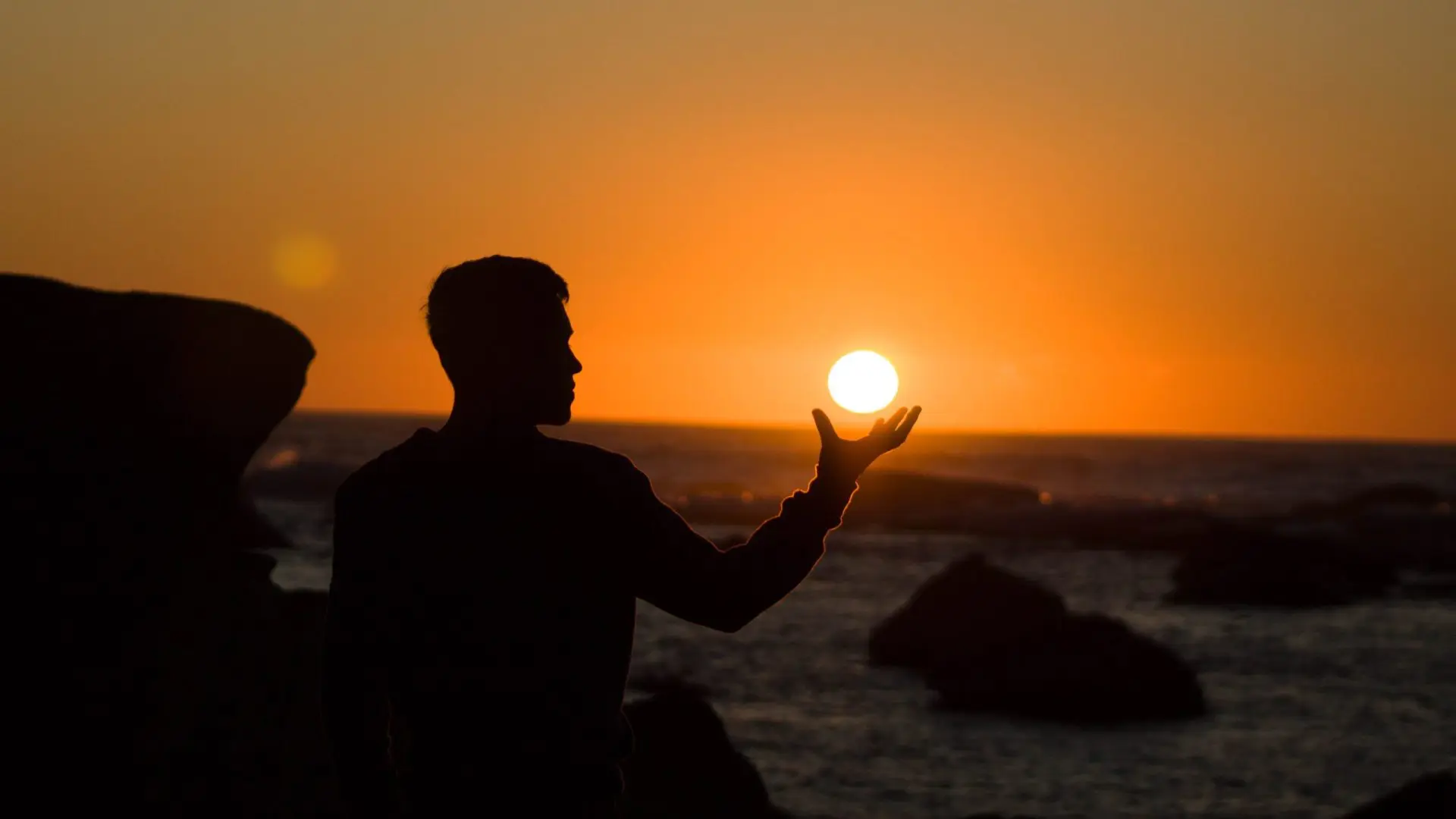Mann sitzt vor Sonnenuntergang am Meer und hält die Sonne